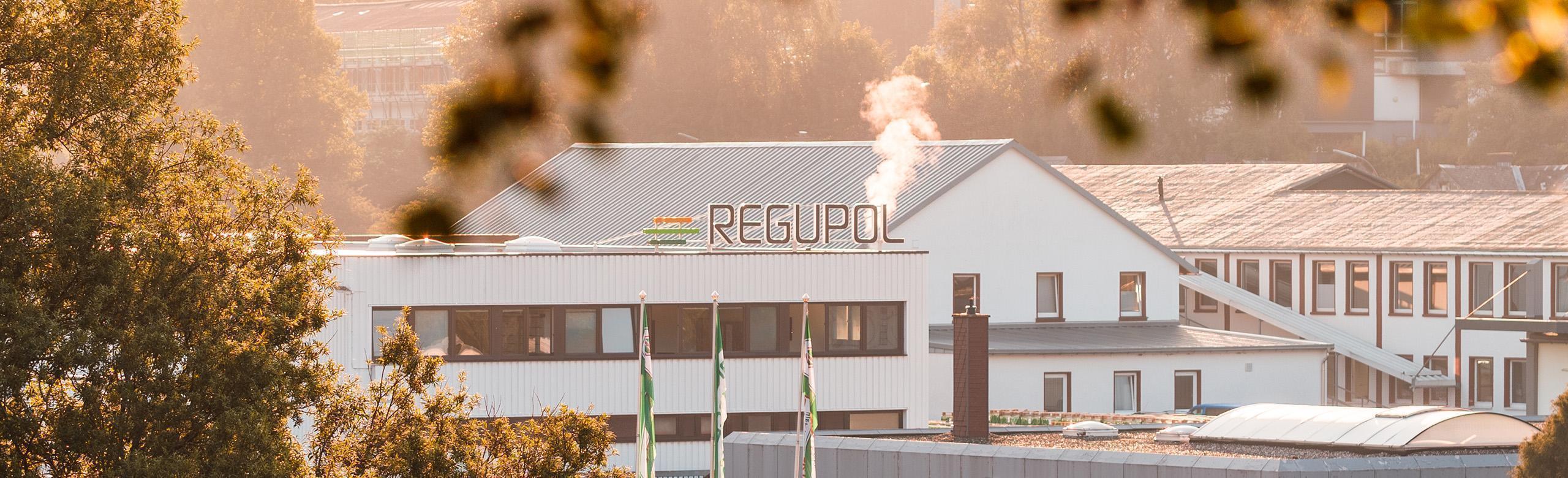 REGUPOL production site at sunrise – facility, sustainability, industry, eco Exterior view of a REGUPOL production site in early morning light. The white industrial building displays the REGUPOL logo on the roof, with steam rising from a chimney. Trees and green REGUPOL flags appear in the foreground.